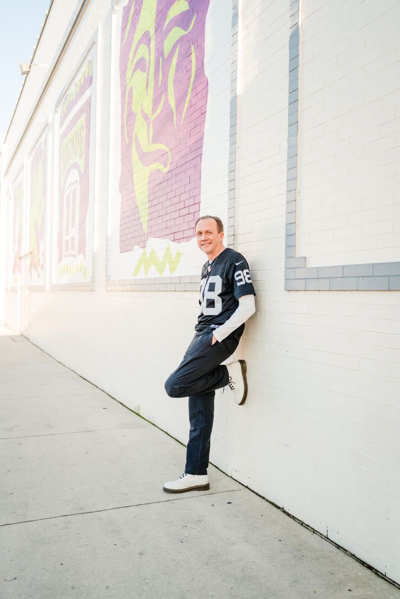 Chad, owner of TTA Fabrication, leaning against a painted brick wall in downtown Fort Myers wearing a Las Vegas Raiders jersey